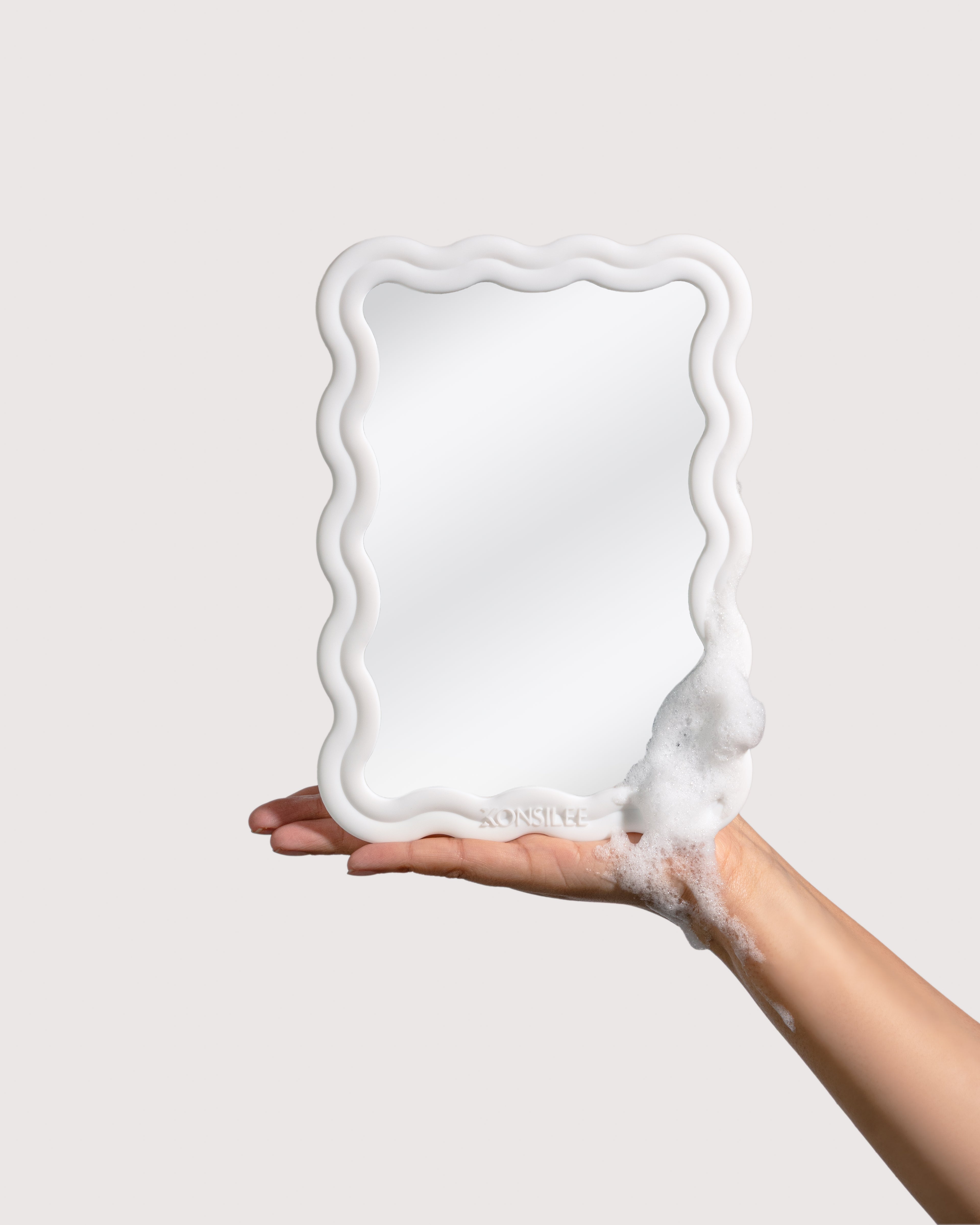A bright-white, shower mirror with a wavy, rectangle frame, held in a person’s soapy hand. The mirror has an embossed Konsilee logo centred at the bottom of the mirror's frame. The background is a solid off-white shade.