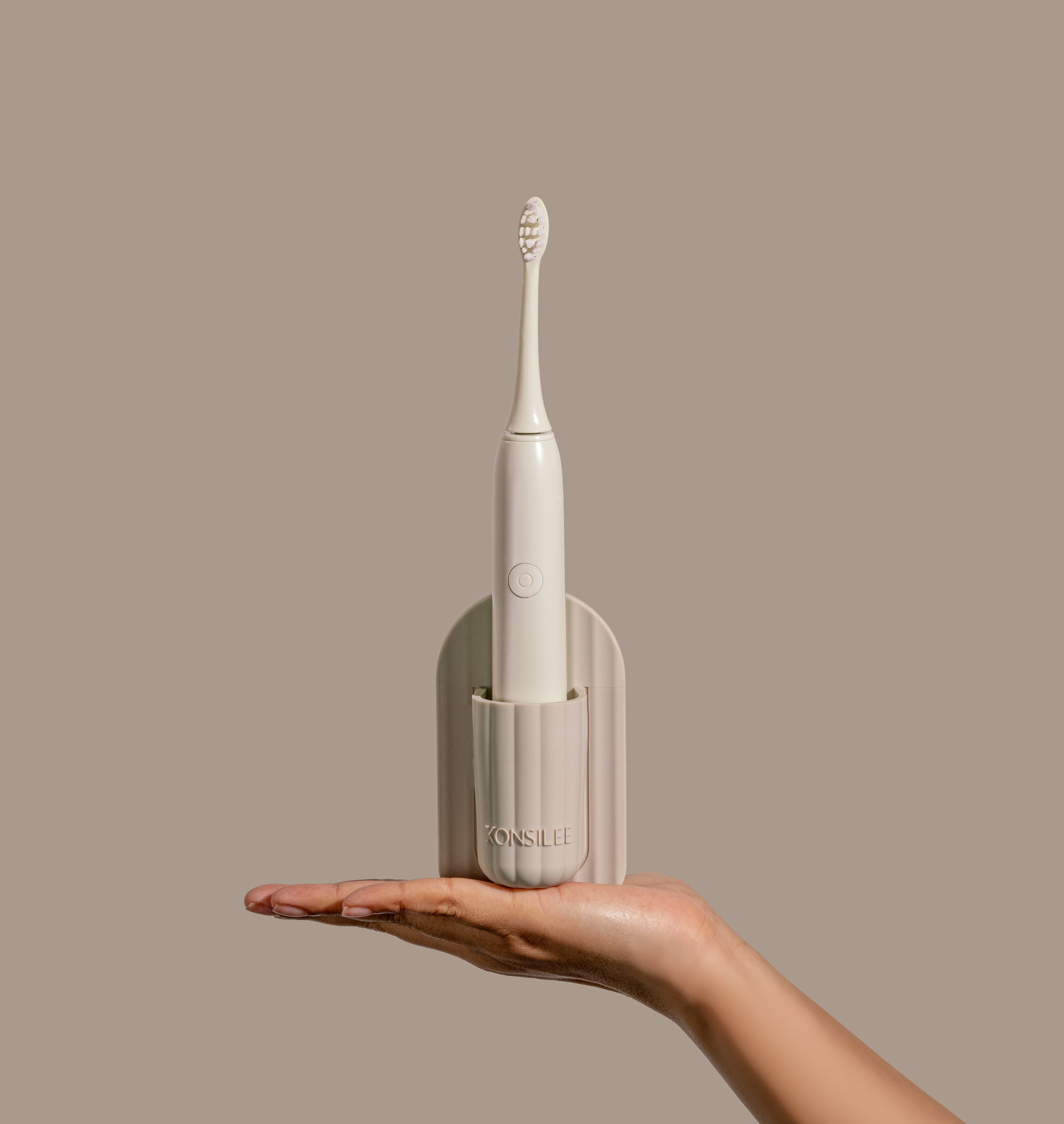 An arched, soft-brown silicone electric toothbrush holder with fluted texture and embossed Konsilee logo. Held in a person's hand against a solid, light-brown backdrop.